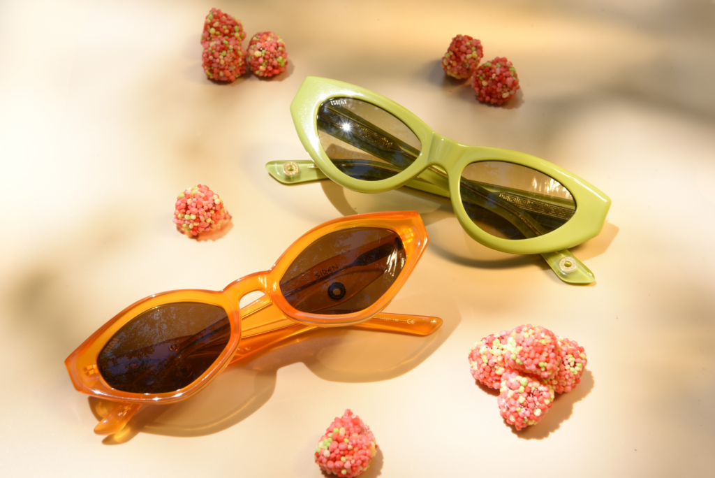 Lime green and orange cat-eye sunglasses surrounded by pink candy on a soft peach background