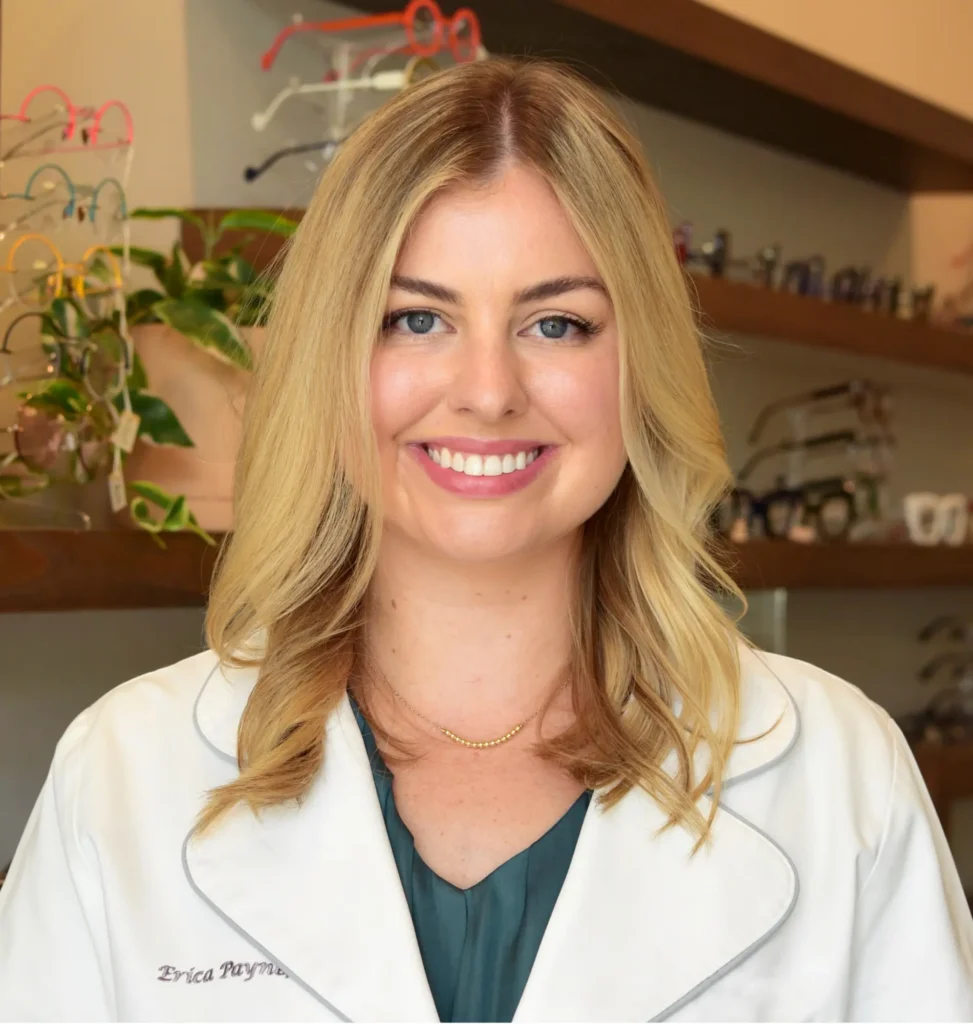 Dr. Erica Payne, optometrist, smiling in a white coat in front of the frame wall