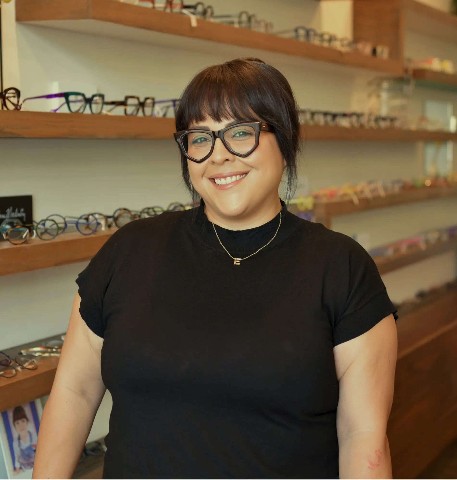 Eden, optician, smiling in black-framed glasses in front of the frame wall