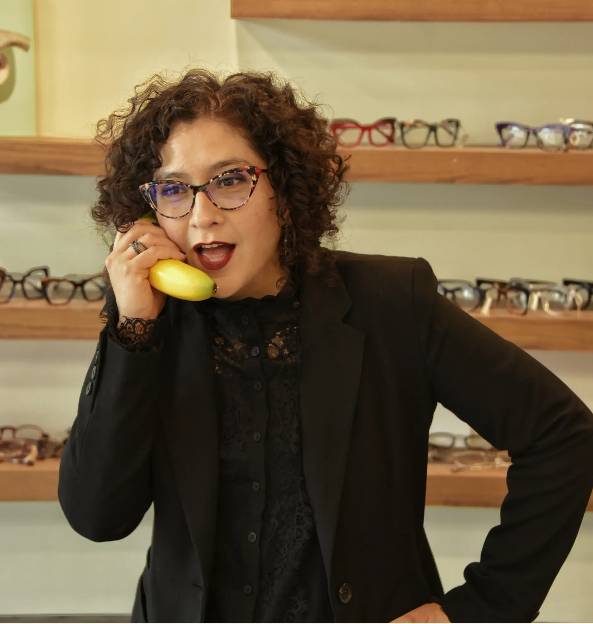 Jessica, optometric technician, holding a banana to her ear like a phone in front of the frame wall