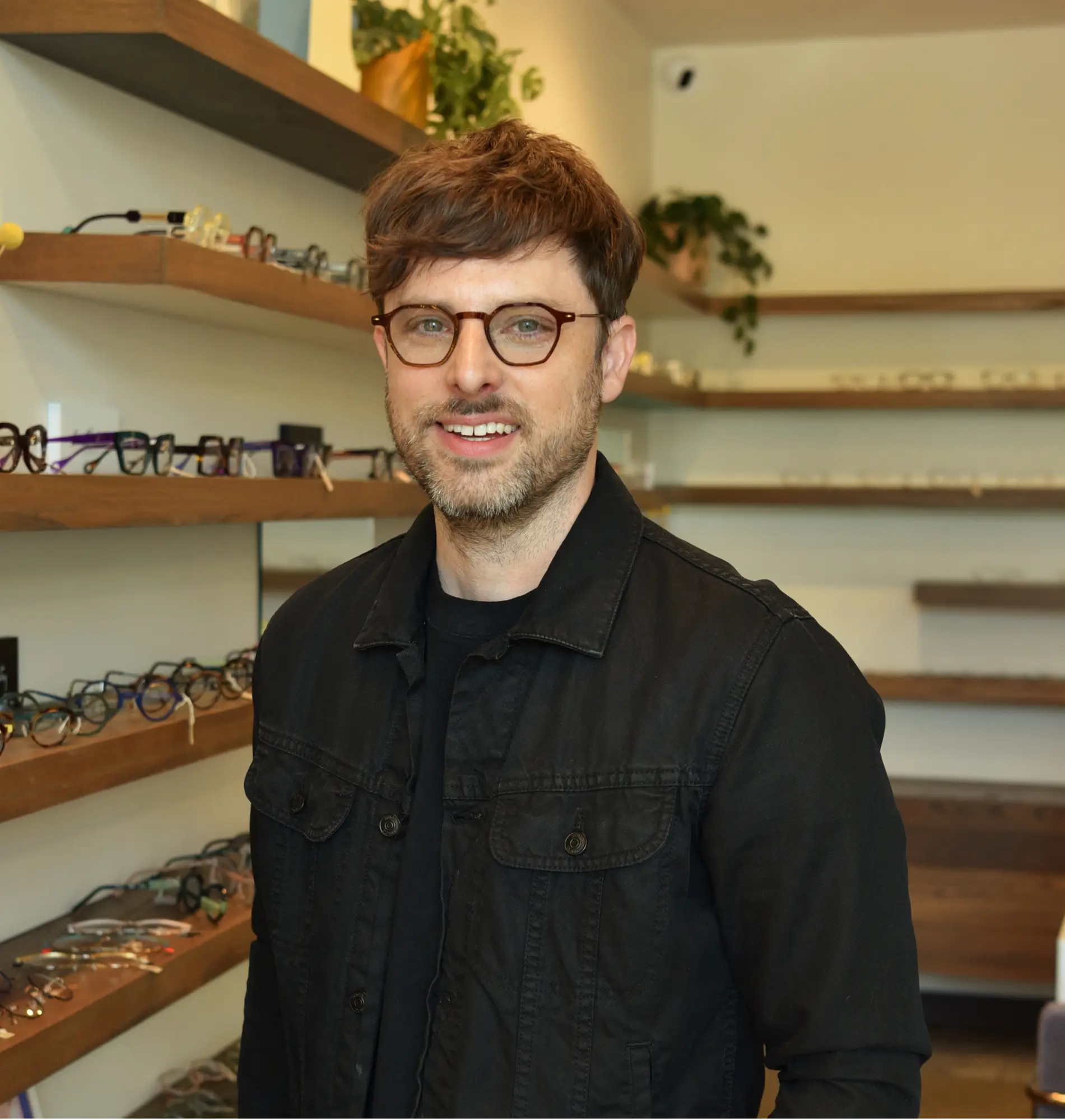 Justin, optical manager, in round glasses and a black jacket in front of the frame wall
