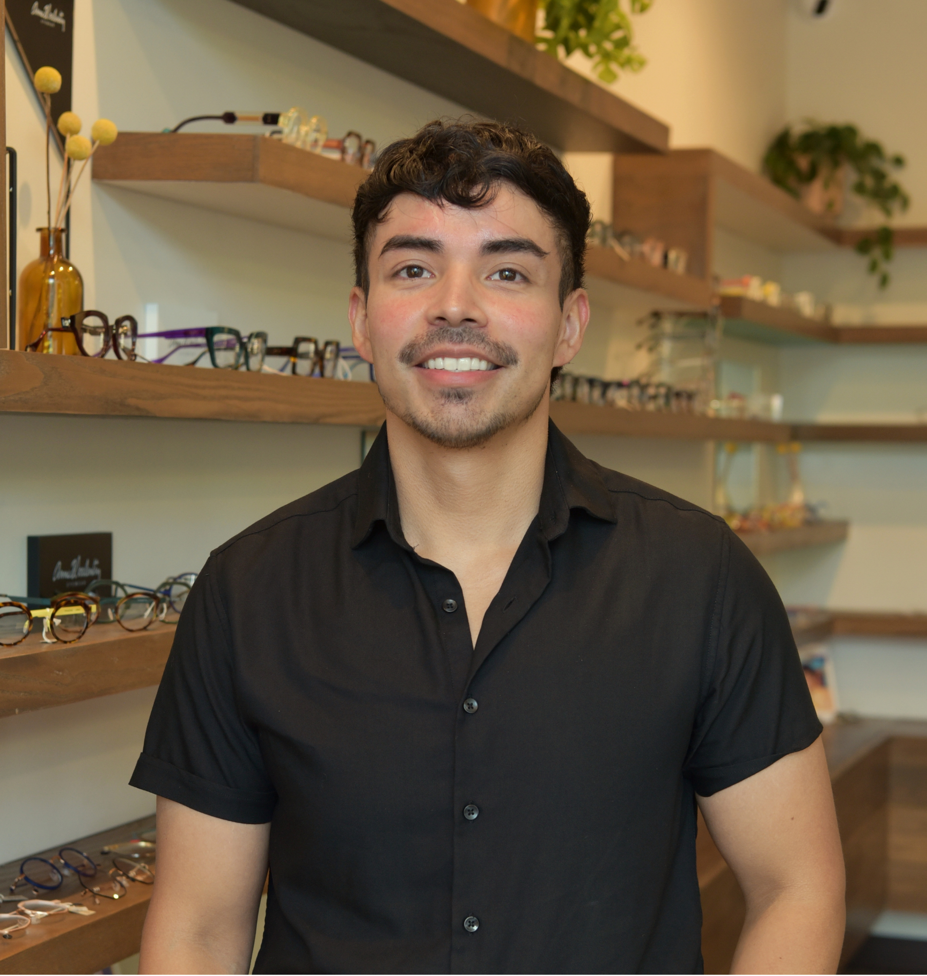 Ruben, optician, smiling in a black button-up shirt in front of the frame wall