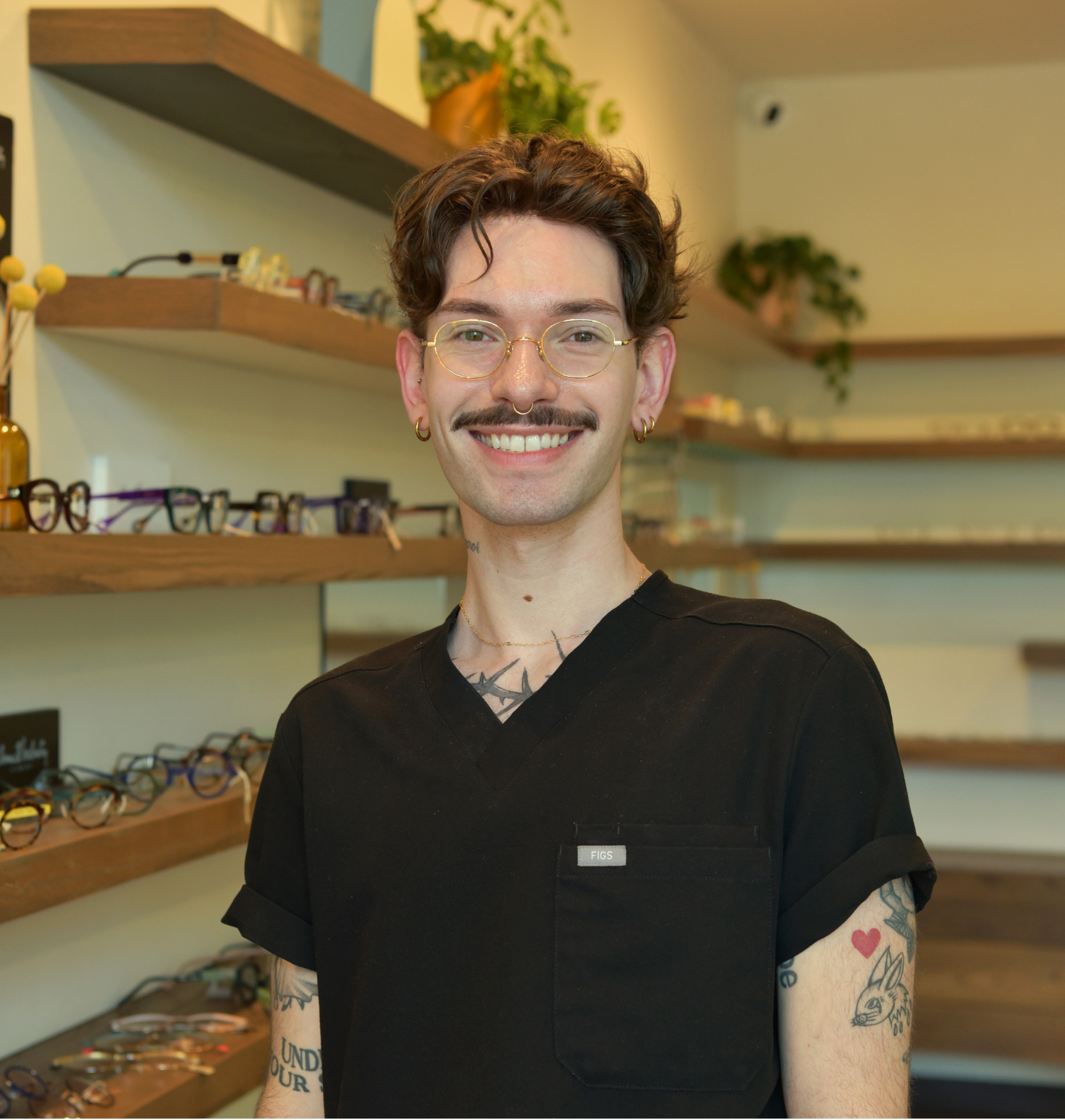 Tyler, optometric technician, in glasses and black scrubs in front of the frame wall