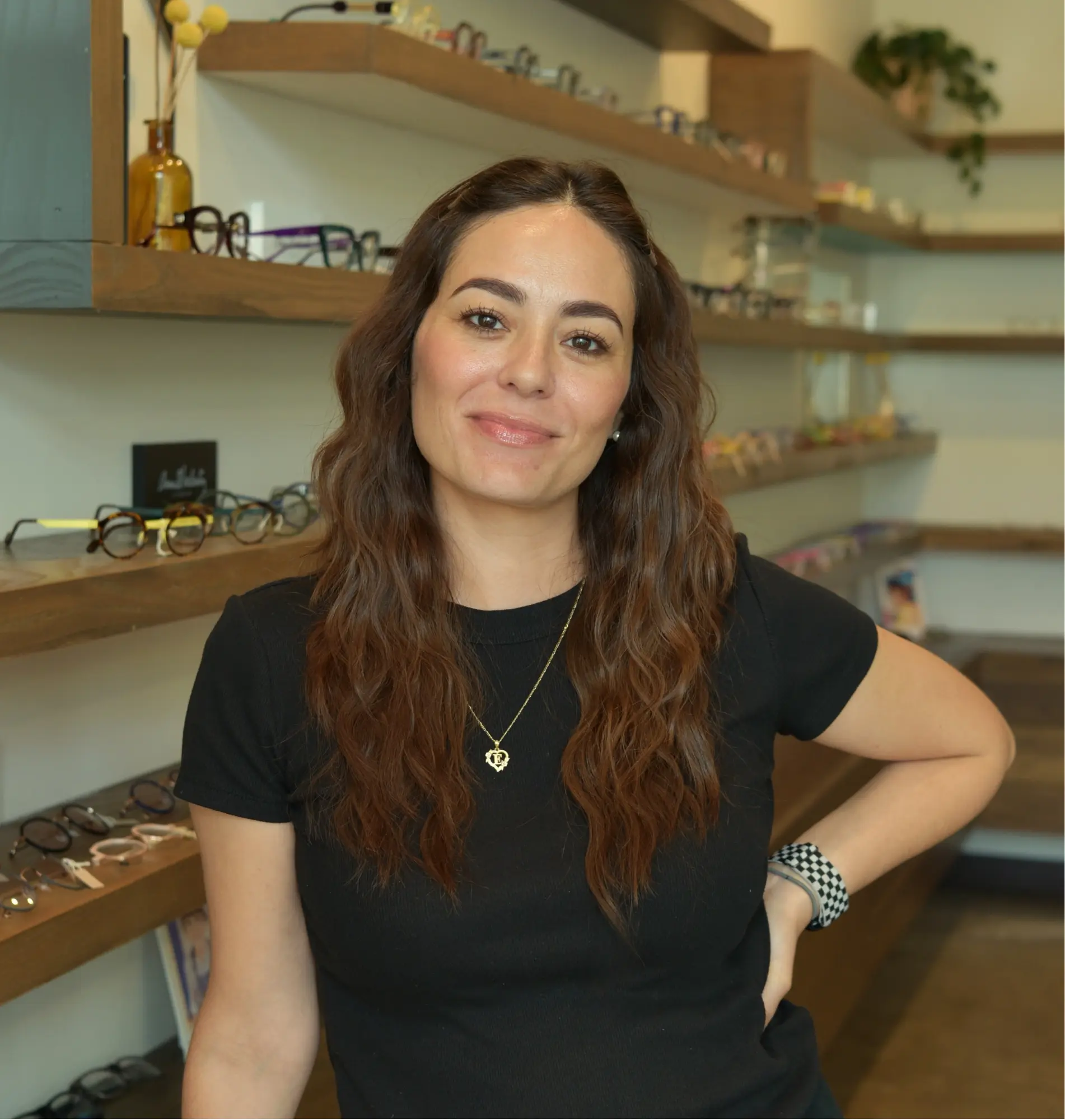 Valerie, optician, smiling with a hand on her hip in front of the frame wall