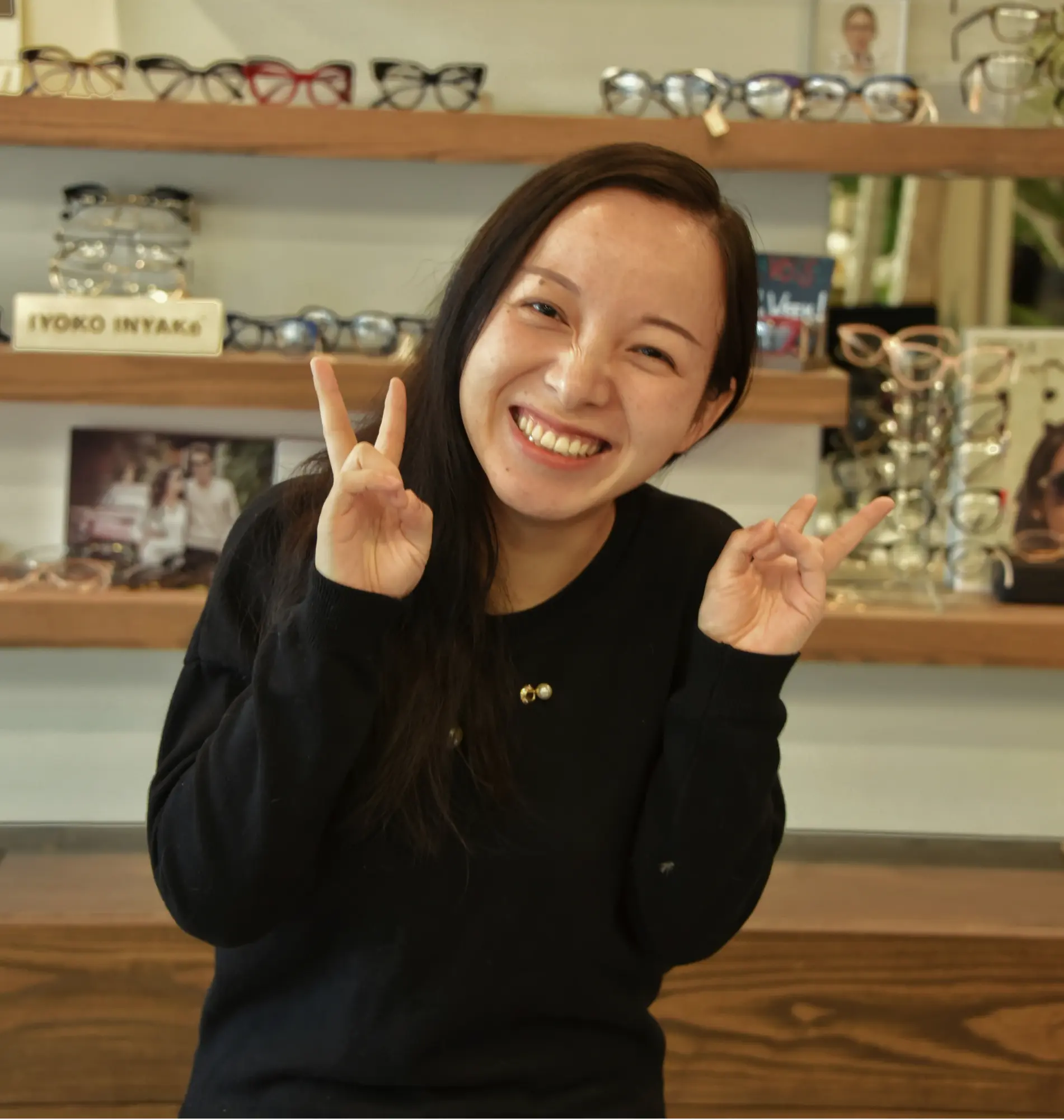 Ying, operations manager and optician, smiling and flashing peace signs in front of the frame wall