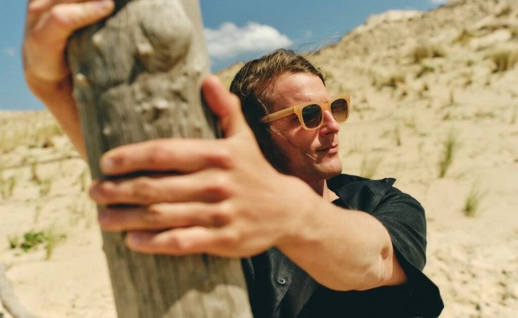 Man in SALT. honey-tinted sunglasses leaning on a wooden post on a sandy hillside